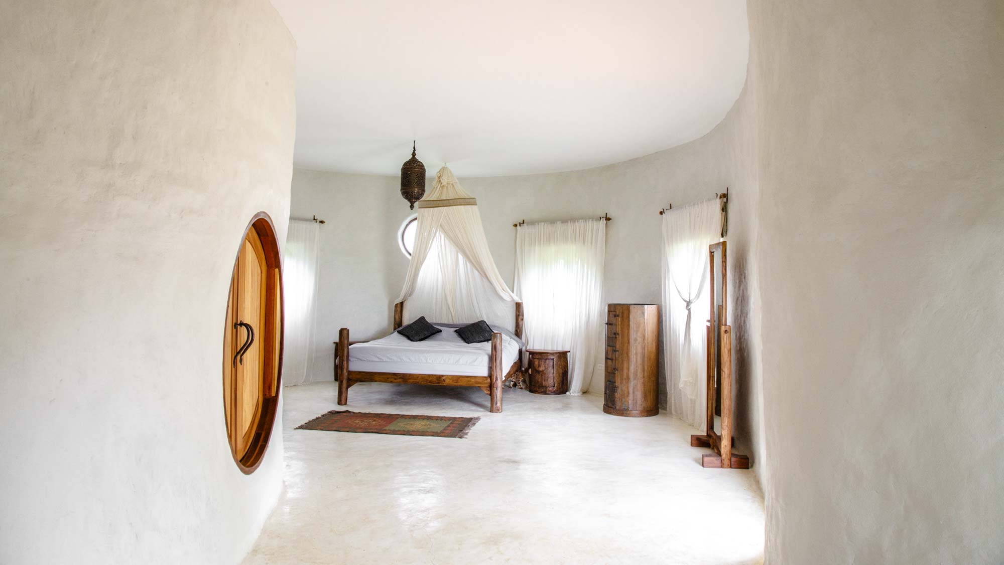 View along a curved adobe walls and round wooden doorway of the bed in the Hobbit Villa at the Chiang Mai Mala Dhara Eco Resort & Yoga Retreat Center, adorned with a hand made cotton mosquito net and surrounded by carefully chosen wooden furniture.