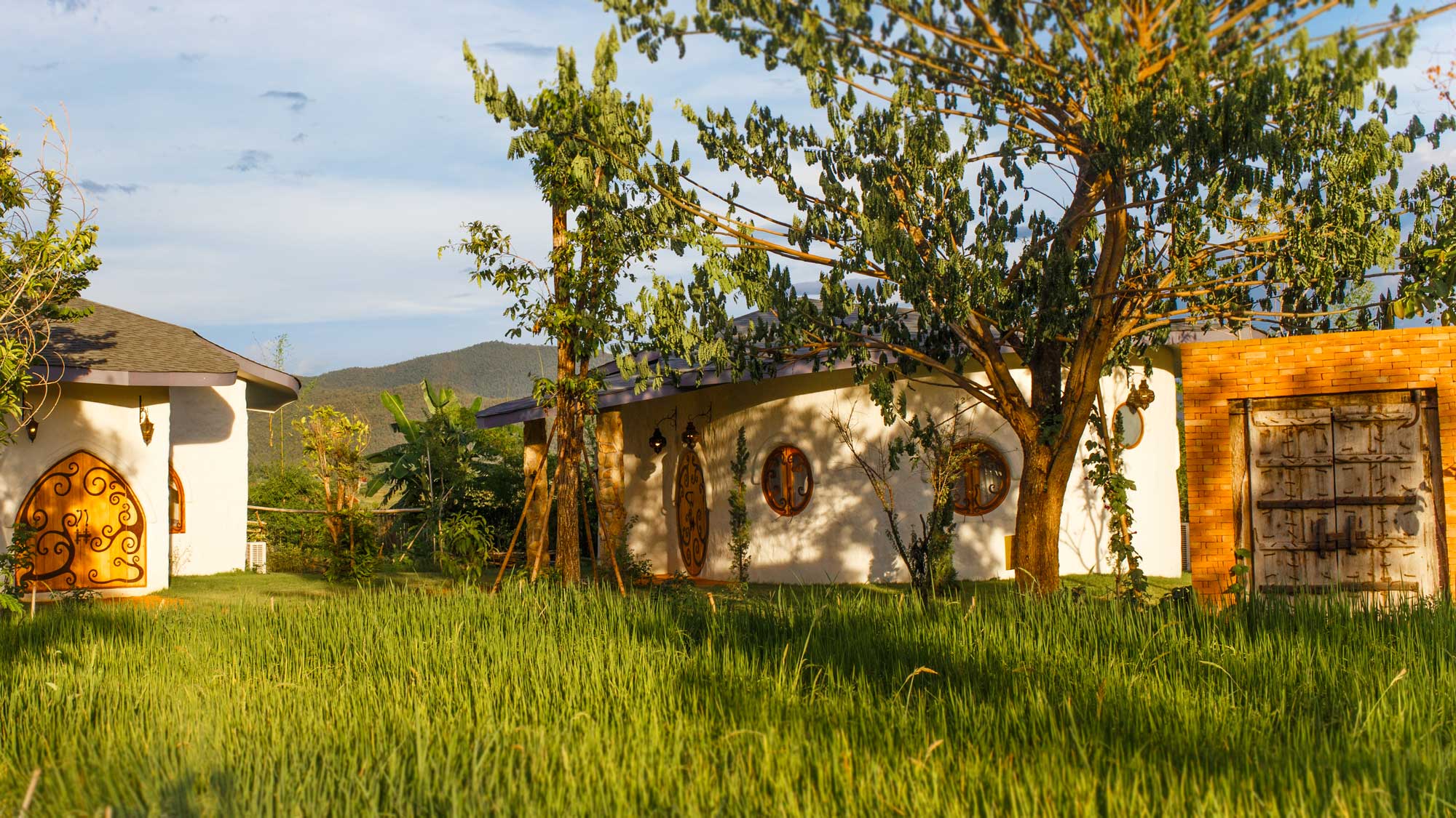 View of two adobe villas and a free standing garden doorway at the Chiang Mai Mala Dhara Yoga Retreat Center & Eco Resort while looking across a small organic rice paddy in the foreground during the late afternoon sunshine.